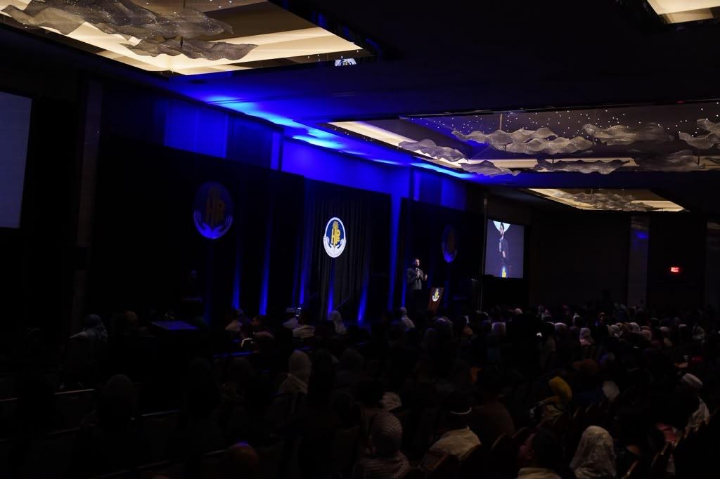 Speaker, projection screen, and audience in a blue-lit ballroom